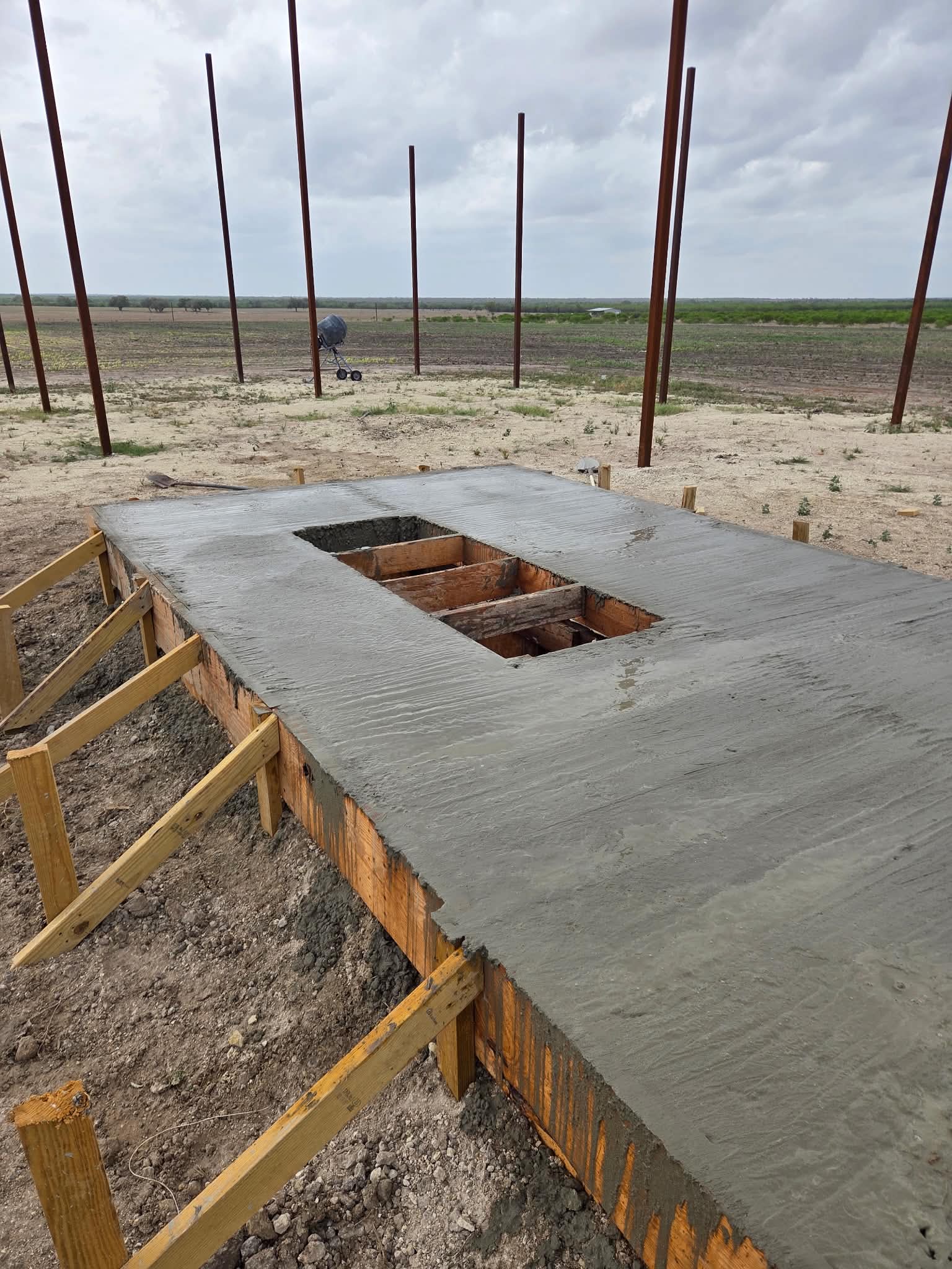 A freshly poured concrete slab with a framed opening through the middle and vertical steel posts set around it, open ranch land behind