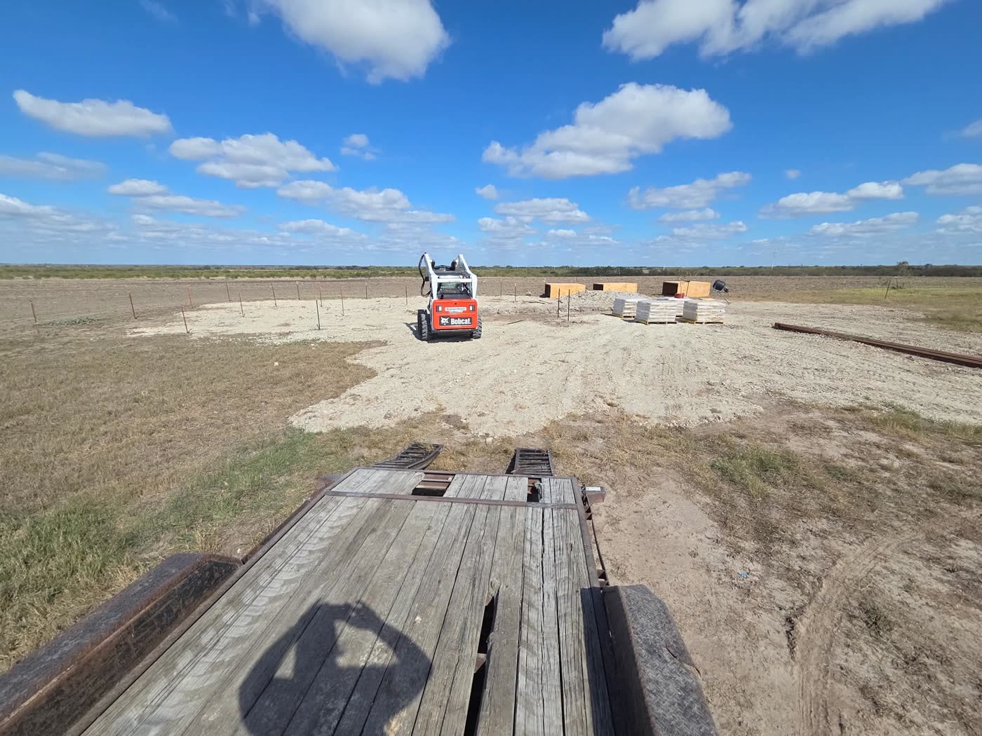 Skid steer cutting a building pad out of open ranch land, stakes set around the perimeter and materials staged on pallets